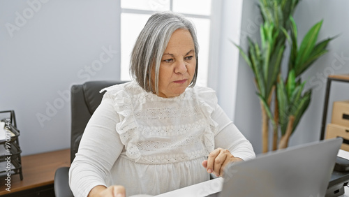 A focused senior woman works diligently in a modern office, surrounded by technology and indoor greenery.