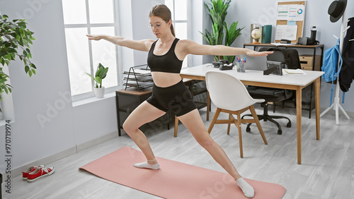 A young adult woman practicing yoga in a modern office setting, dressed in workout attire, creating a scene of work-life balance.