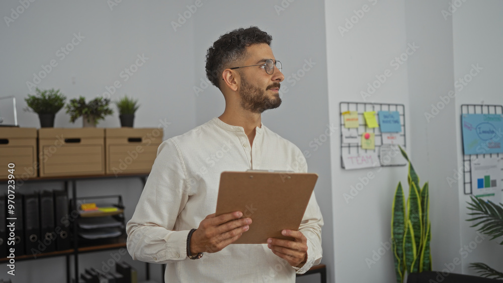 A handsome young hispanic man in light-colored clothing holds a clipboard while standing in a modern office with minimalist decor. A handsome young hispanic man in light-colored clothing holds a clipboard while standing in a modern office with minimalist decor.