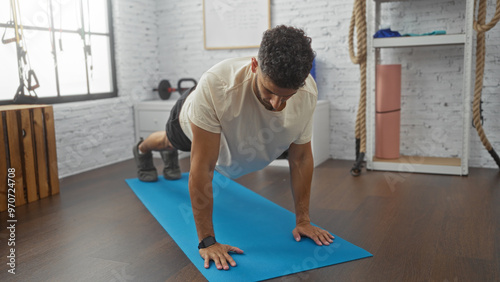 Young, hispanic man doing a plank exercise on a blue mat in a well-equipped gym with white brick walls.