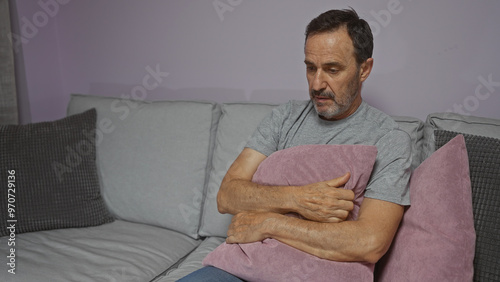 Obraz na plátně Elderly man in a living room indoors, holding a pillow and looking pensive, seated on a comfortable sofa with a neutral expression, implying a sense of introspection or solitude