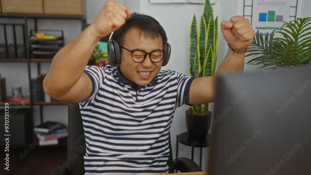 Young chinese man celebrates in an office room with headphones. Young chinese man celebrates in an office room with headphones.