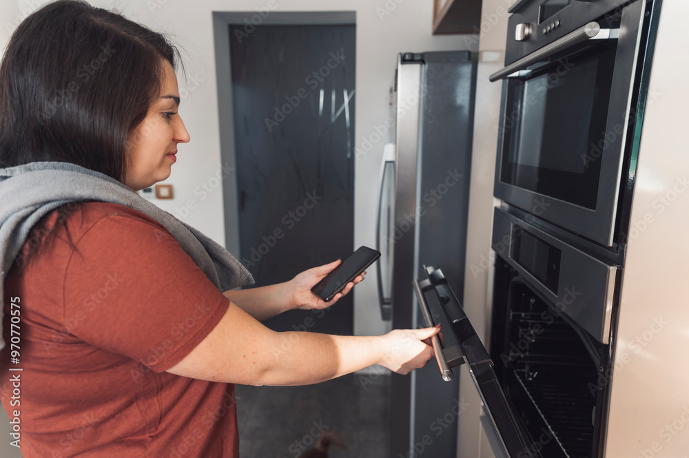 A woman in a modern kitchen is opening a drawer while holding a tablet. The kitchen features wooden accents and stainless steel appliances, with natural light coming in. A woman in a modern kitchen is opening a drawer while holding a tablet. The kitchen features wooden accents and stainless steel appliances, with natural light coming in.