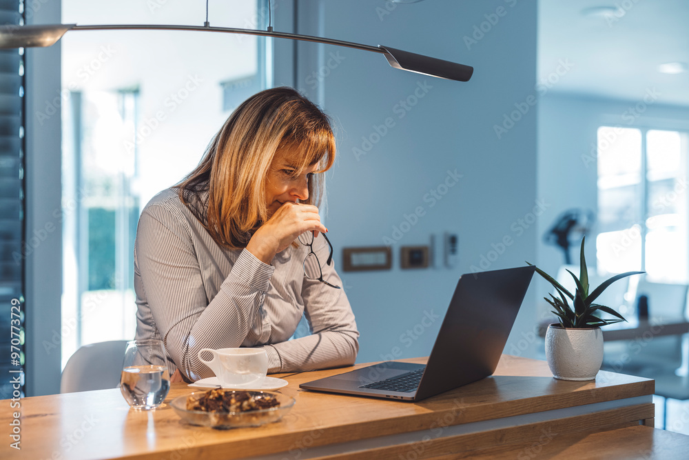 Obraz premium A woman sitting at a wooden table, looking at her laptop with a thoughtful expression. She has a cup of coffee and a glass of water beside her, with a small plant in the background. The setting is bri