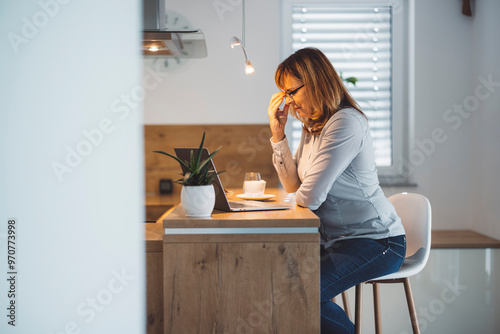 A woman sitting at a kitchen island, looking stressed while using a laptop. She has long brown hair and wears a light blue shirt. A small plant and a cup are on the counter.