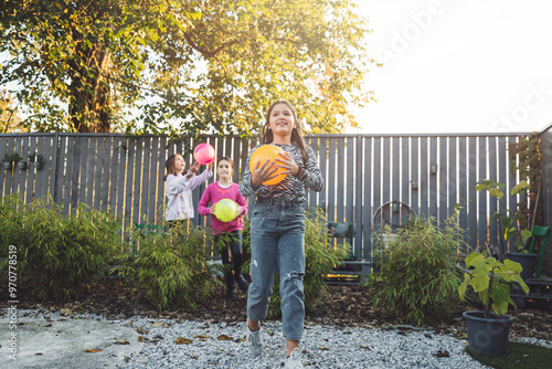 A group of children playing with colorful balls in a sunny backyard. One girl in the foreground holds an orange ball, while two others in the background are playing with pink and yellow balls. The set