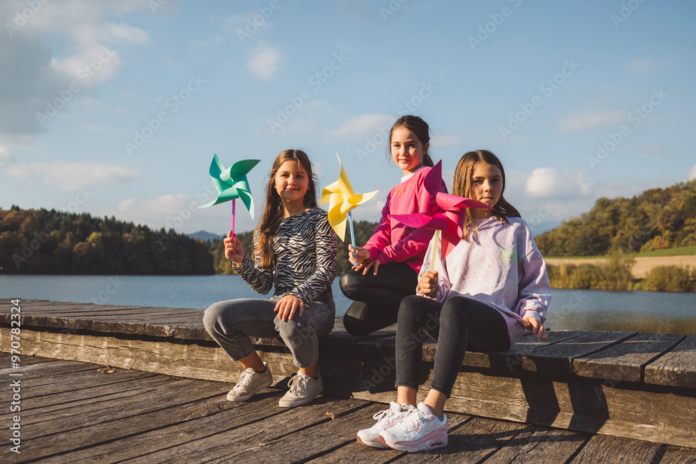 Fototapeta premium Three children sitting on a wooden dock by a lake, holding colorful pinwheels. They are smiling and enjoying a sunny day with a scenic background of trees and blue sky.