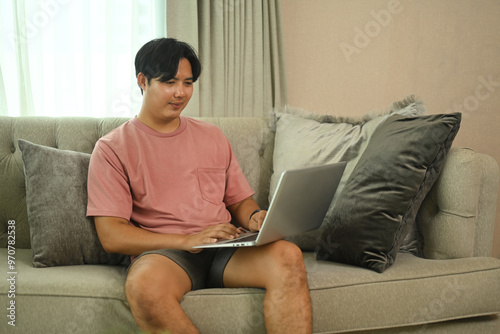 Smiling young asian man in casual clothing using laptop on comfortable sofa at home