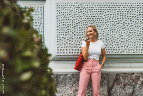 A woman stands against a textured wall, smiling while holding a phone. She wears a white t-shirt and pink high-waisted pants, complemented by a red handbag. Lush greenery is partially visible in the f