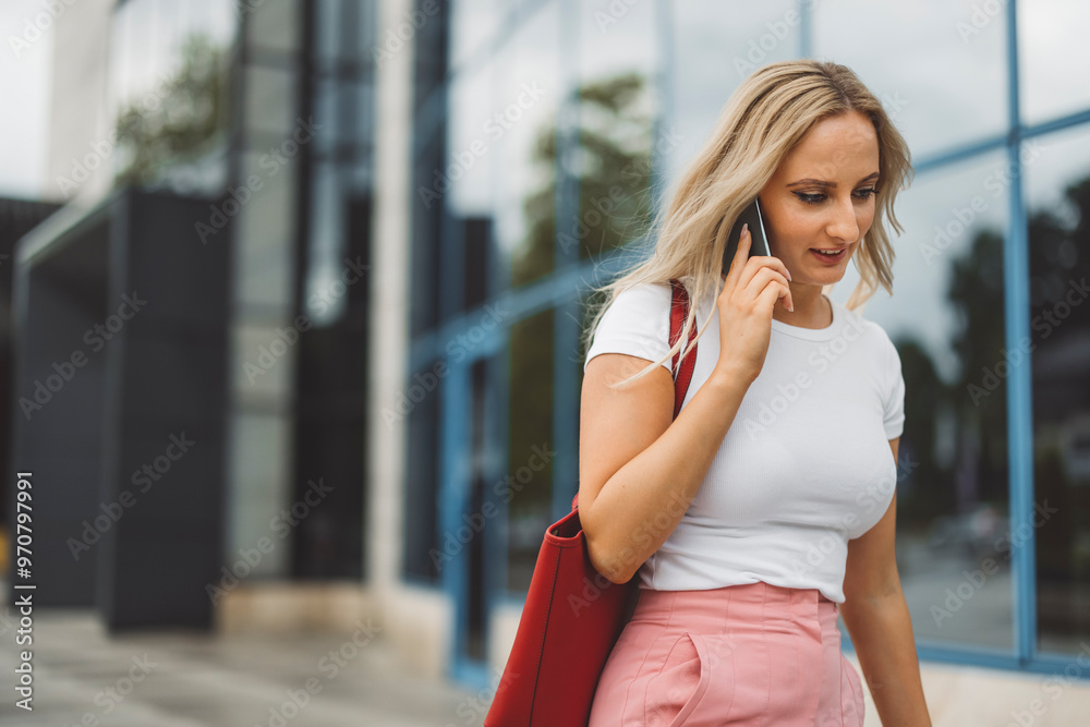 A young woman walking outside a modern building while talking on her smartphone. She has long blonde hair and wears a white t-shirt paired with pink trousers. A red handbag is slung over her shoulder, A young woman walking outside a modern building while talking on her smartphone. She has long blonde hair and wears a white t-shirt paired with pink trousers. A red handbag is slung over her shoulder,