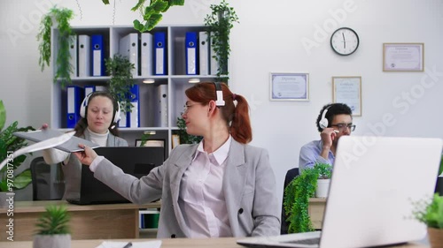 relationship at work, woman with headset and glasses works in call center and gives documents to female colleague sitting at desk in office photo