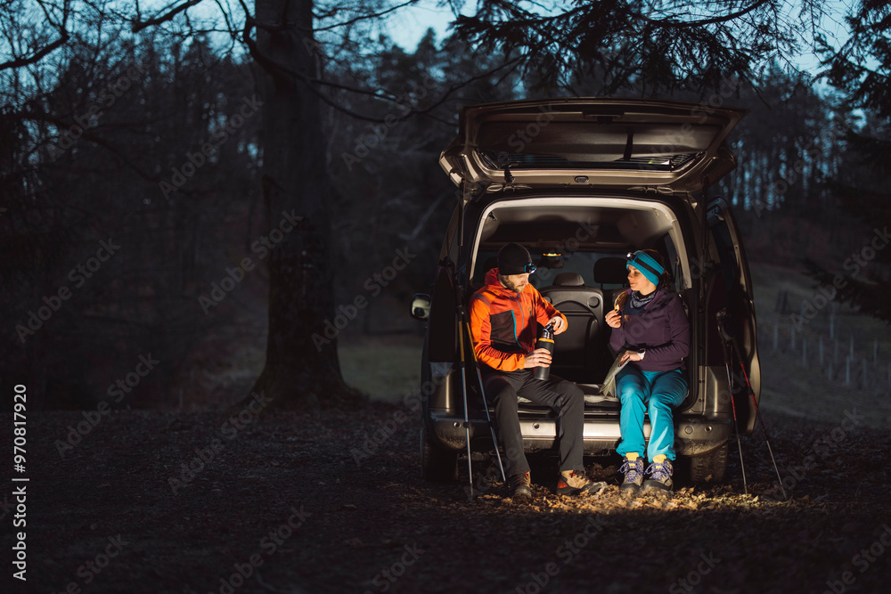 A couple enjoying a cozy evening by their van in the woods, illuminated by soft light. Camping in nature. They are sitting on camping chairs, sharing snacks and laughter, surrounded by trees. A couple enjoying a cozy evening by their van in the woods, illuminated by soft light. Camping in nature. They are sitting on camping chairs, sharing snacks and laughter, surrounded by trees.