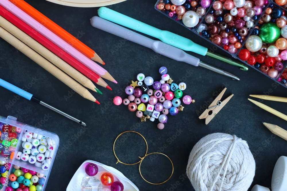 Colorful beads, coloring pencils, glitter, needles, string, hoop, tape and scissors on dark background. Various colorful craft supplies. Flat lay.