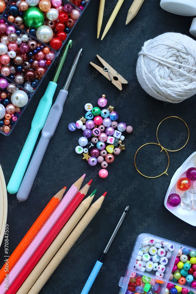 Colorful beads, coloring pencils, glitter, needles, string, hoop, tape and scissors on dark background. Various colorful craft supplies. Flat lay.