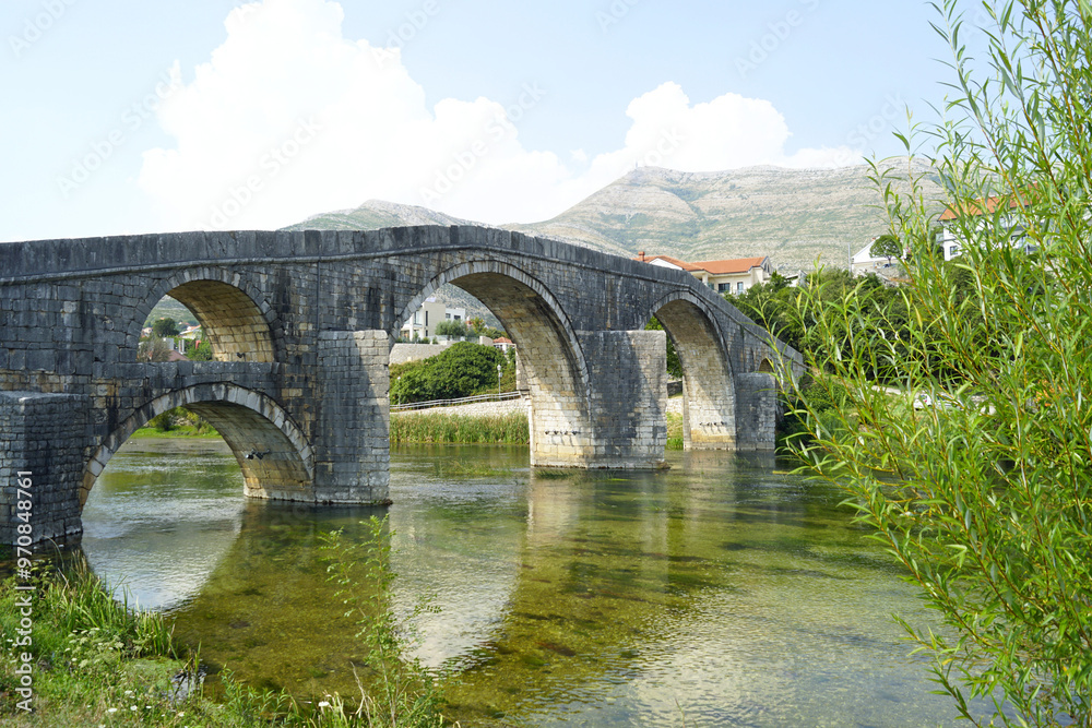 Summer landscape with Arslanagić bridge: view of the old high bridge with arches built over the clear waters of the Trebišnjica river. Architectural heritage of Trebinje, Bosnia and Herzegovina. Summer landscape with Arslanagić bridge: view of the old high bridge with arches built over the clear waters of the Trebišnjica river. Architectural heritage of Trebinje, Bosnia and Herzegovina.
