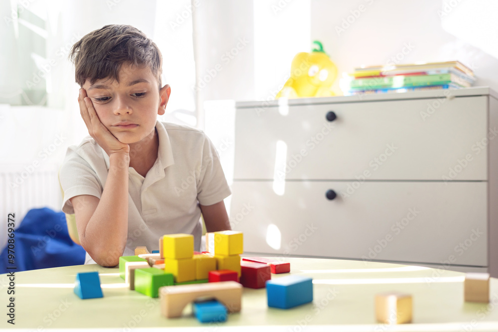 Little boy sad and tired plays with wooden blocks at children's table in bright room. focus on mental health and the role of speech therapy, psychological support and educational play. Little boy sad and tired plays with wooden blocks at children's table in bright room. focus on mental health and the role of speech therapy, psychological support and educational play.