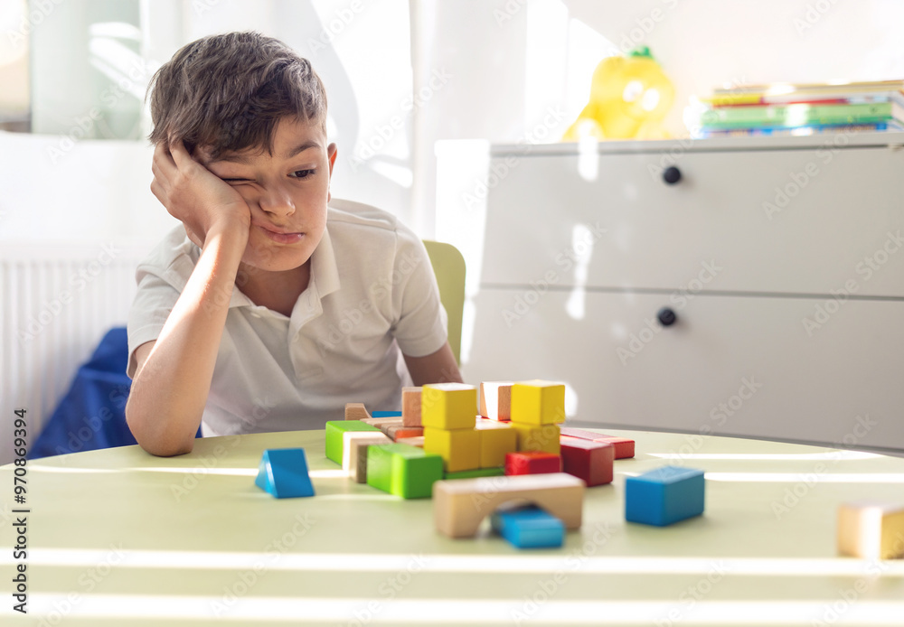 Little boy sad and tired plays with wooden blocks at children's table in bright room. focus on mental health and the role of speech therapy, psychological support and educational play. Little boy sad and tired plays with wooden blocks at children's table in bright room. focus on mental health and the role of speech therapy, psychological support and educational play.