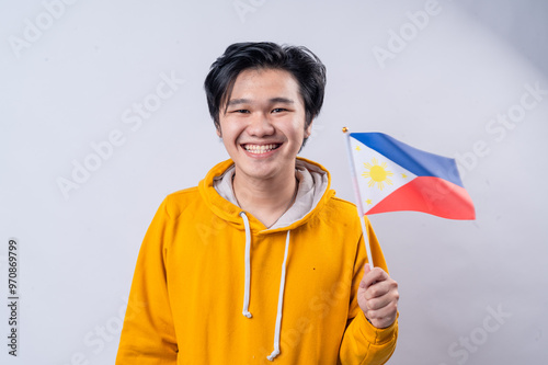Obraz na plátně An Asian man in a yellow hoodie stands against a white background, smiling widely while holding the Philippine flag