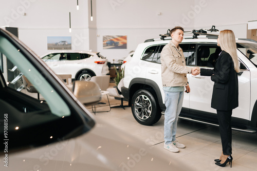 Remote wide shot of happy male car owner getting key from smiling female auto dealer at dealership. Handsome young male taking key to bought car and sealing deal with handshake in auto showroom