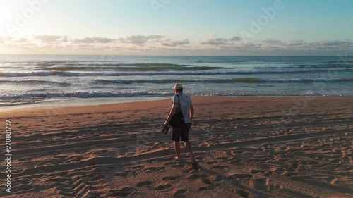 A man strolls on the beach at sunrise, carrying an orange umbrella and a black bag, wearing a white hat, light shirt, shorts, and flip-flops. Slow motion. photo