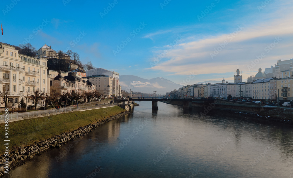 Obraz premium A serene winter morning in Salzburg with snow-covered buildings and a peaceful river promenade