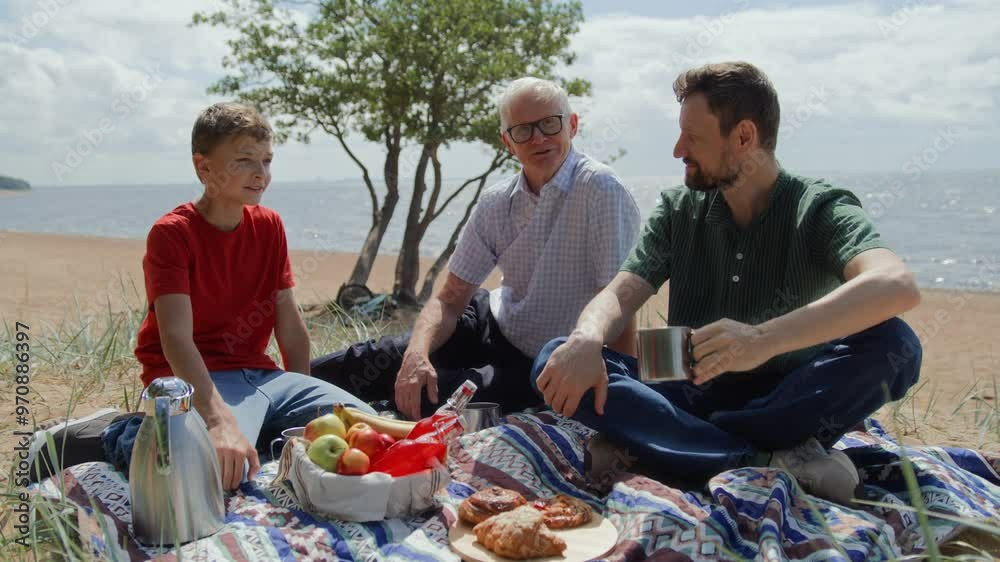 Boy, father and grandfather relaxing on beach having picnic on blanket. Grandson, adult father and older man, sitting, chatting and enjoying buns and tea from thermos. Male generations togetherness