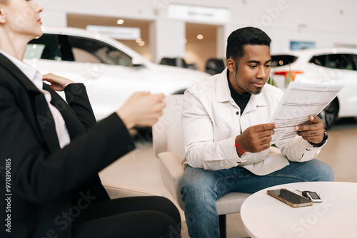 Focused African-American male buyer carefully reading car purchase agreement before signing in dealership office. Positive saleswoman car dealer consulting doubtful buyer at showroom.