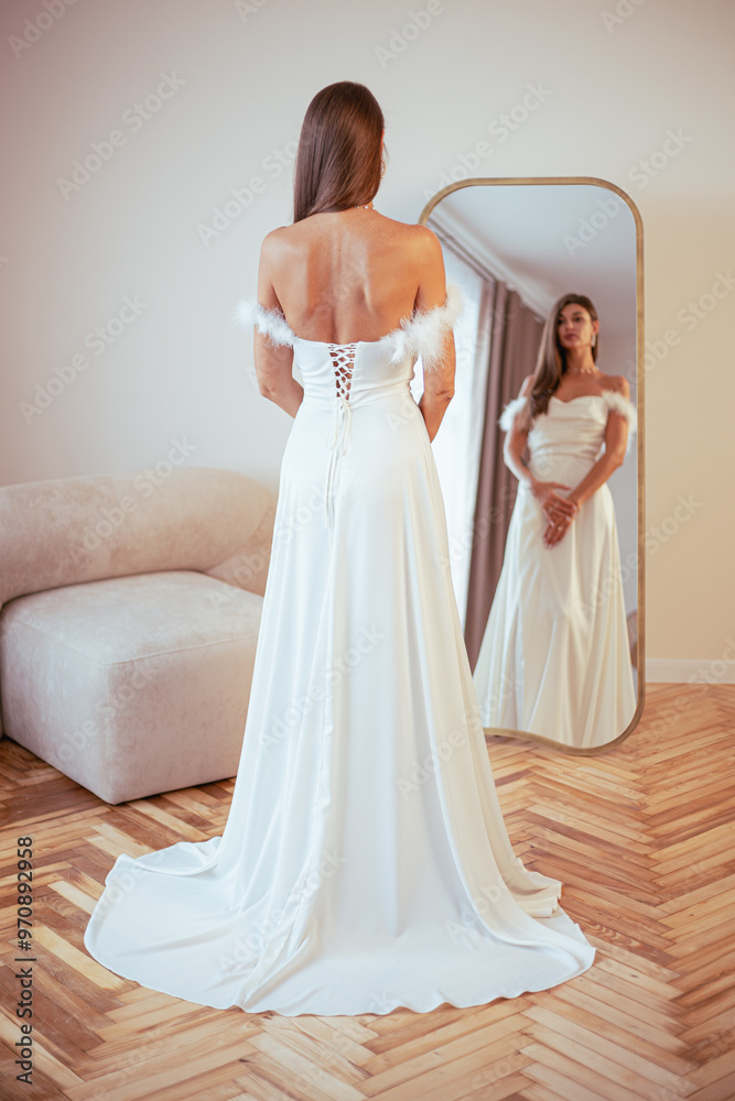 Fototapeta premium Beautiful brunette woman in a white dress. Stands in the room in front of the mirror, looks at her reflection and admires herself