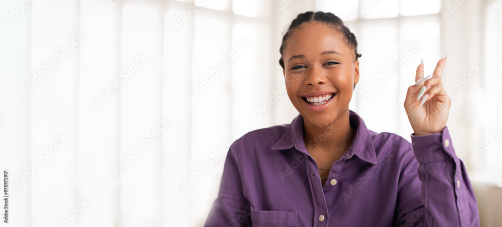 A young black woman happily smiles and raises her hand in a playful gesture indoors, surrounded by bright natural light streaming through large windows. The atmosphere feels warm and inviting. A young black woman happily smiles and raises her hand in a playful gesture indoors, surrounded by bright natural light streaming through large windows. The atmosphere feels warm and inviting.