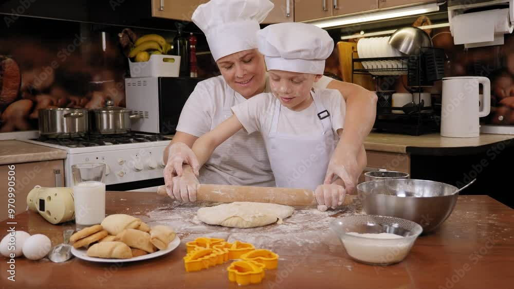 Close-up of a little curious boy in a white apron and a cap rolls out the dough with a rolling pin at home in the kitchen, his loving mother is sitting next to him. Close-up of a little curious boy in a white apron and a cap rolls out the dough with a rolling pin at home in the kitchen, his loving mother is sitting next to him.