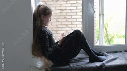 A teenage girl with long hair sits on the windowsill and reads on a tablet. A smart, modern kid is engaged in self-development, reading a lot of literature on the gadget