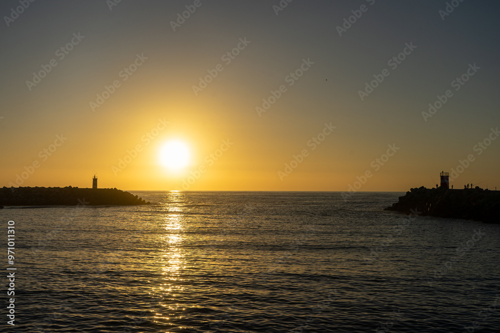 A breathtaking sunset at Nazaré, Portugal, with the sun dipping below the horizon, framed perfectly between two lighthouses at the entrance of the marina, casting golden reflections on the sea. A breathtaking sunset at Nazaré, Portugal, with the sun dipping below the horizon, framed perfectly between two lighthouses at the entrance of the marina, casting golden reflections on the sea.