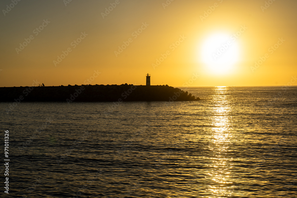 A breathtaking sunset at Nazaré, Portugal, with the sun dipping below the horizon, framed perfectly between two lighthouses at the entrance of the marina, casting golden reflections on the sea. A breathtaking sunset at Nazaré, Portugal, with the sun dipping below the horizon, framed perfectly between two lighthouses at the entrance of the marina, casting golden reflections on the sea.