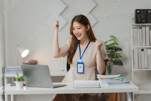 Success Story: A young businesswoman celebrates a significant achievement, her hands raised in triumph as she stares triumphantly at her laptop screen, radiating joy and determination. The image conve photo