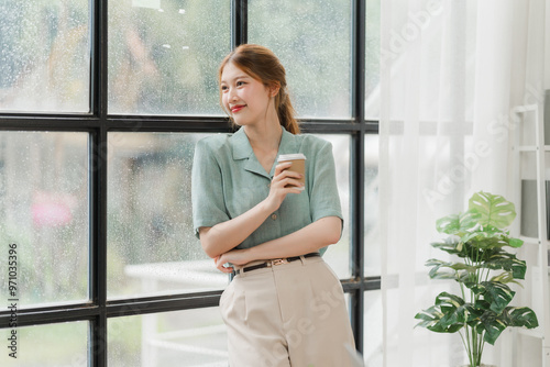 Contemplative Coffee Break: Young woman in stylish office attire enjoys a moment of reflection by the window, coffee in hand, embodying modern work-life balance.