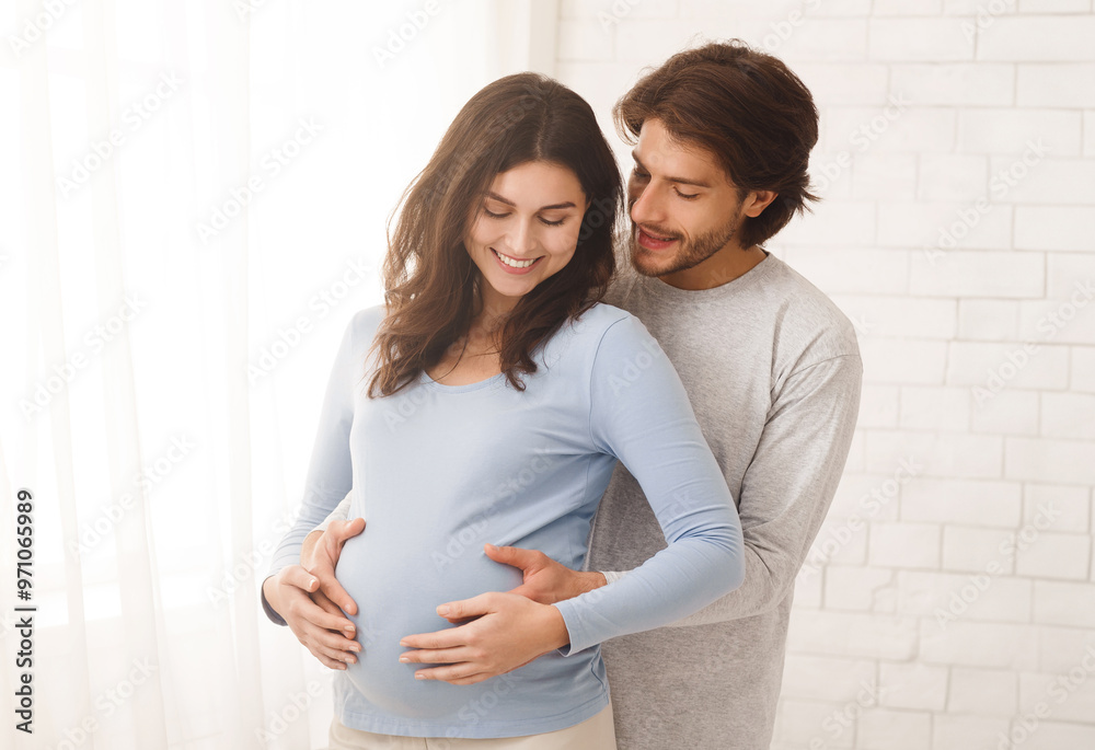 Loving man hugging his pregnant wife from behind standing near window at home, copy space Loving man hugging his pregnant wife from behind standing near window at home, copy space
