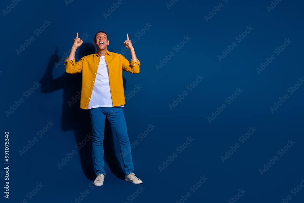 Full length portrait of nice young man indicate fingers up empty space wear yellow shirt isolated on dark blue color background Full length portrait of nice young man indicate fingers up empty space wear yellow shirt isolated on dark blue color background