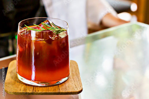 Glass of iced tea carcade or cocktail garnished with rosemary on wooden coaster, placed on reflective glass table with blurred background, creating relaxing atmosphere photo