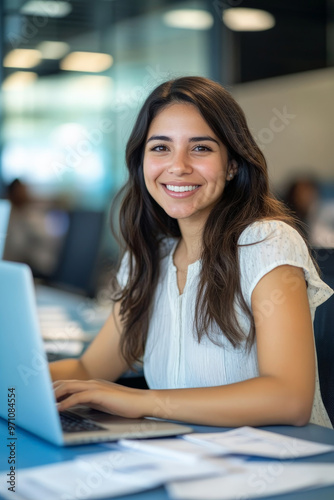 Free Download Young Hispanic female entrepreneur working in a modern corporate office, smiling at the camera, typing on a laptop with documents spread out on her desk, with a blue background. - GFXLoad