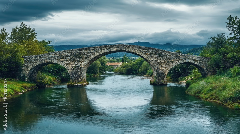An ancient arched stone bridge crossing a river under a cloudy sky. An ancient arched stone bridge crossing a river under a cloudy sky.
