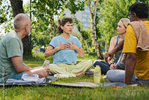 Elderly brunette woman in activewear touching chest while explaining group of diverse people how yoga supporting her health