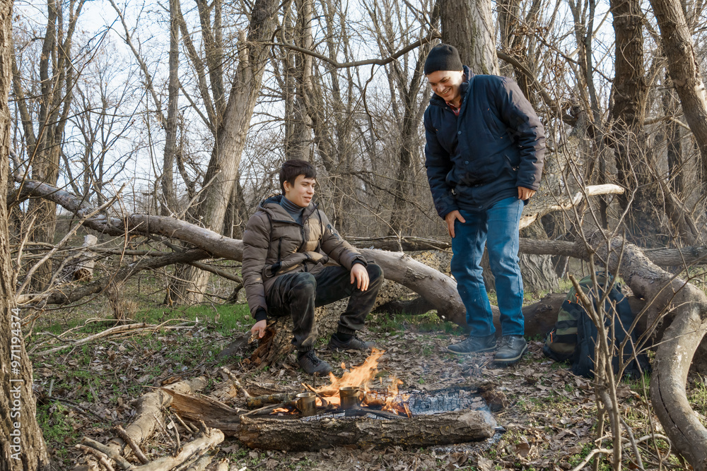 Two men make a campfire for cooking in the forest, father and son sitting on a log while hiking and outdoor activities, early spring landscape