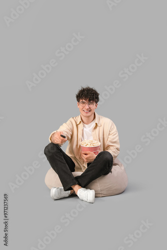 Young man in eyeglasses sitting on beanbag chair with popcorn and remote control on grey background