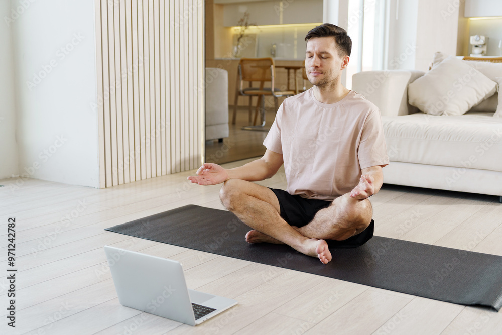 A man meditates in a serene living room, seated on a yoga mat, with a laptop placed in front of him. A man meditates in a serene living room, seated on a yoga mat, with a laptop placed in front of him.