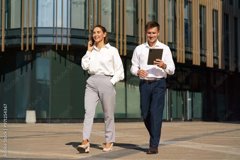 Fototapeta premium A man and woman in business attire walk confidently outdoors, with the woman on a phone call and the man holding a tablet.