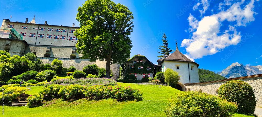Naklejka premium Burg Hohenwerfen in Austria is beautifully nestled in lush greenery, with the majestic Alps in the background. This medieval castle offers a picturesque view of history, nature, and architecture blend