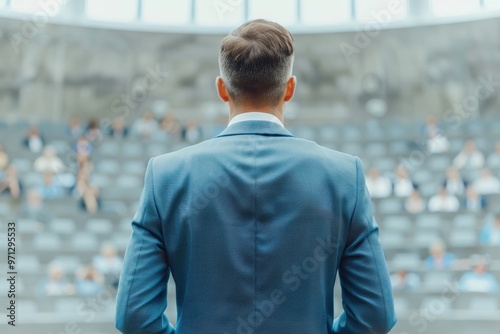 A back view of a man standing and speaking to a large audience in a conference hall, representing public speaking or leadership.