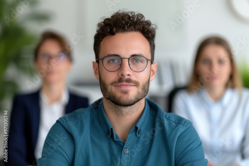 Portrait of a confident young man with glasses in a professional office environment, sitting in front of two colleagues out of focus.