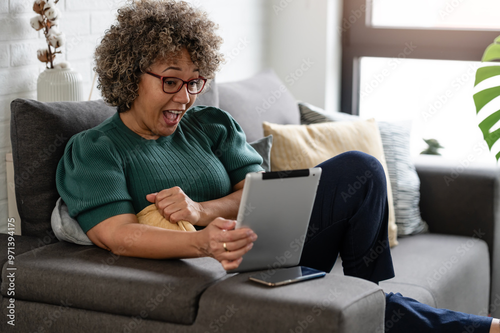 A mature woman with curly gray hair and red glasses sits on a sofa, using a tablet. She is focused and engaged, dressed casually in a green sweater, in a comfortable home setting A mature woman with curly gray hair and red glasses sits on a sofa, using a tablet. She is focused and engaged, dressed casually in a green sweater, in a comfortable home setting