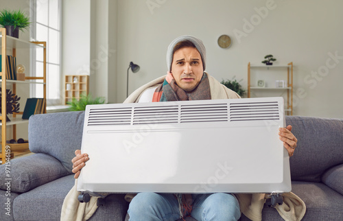 Portrait of a frozen man, bundled in outerwear and wrapped in a blanket, seated on a living room sofa. Holding an electric heater, he confronts the cold, embodying the frozen solitude of the season. photo